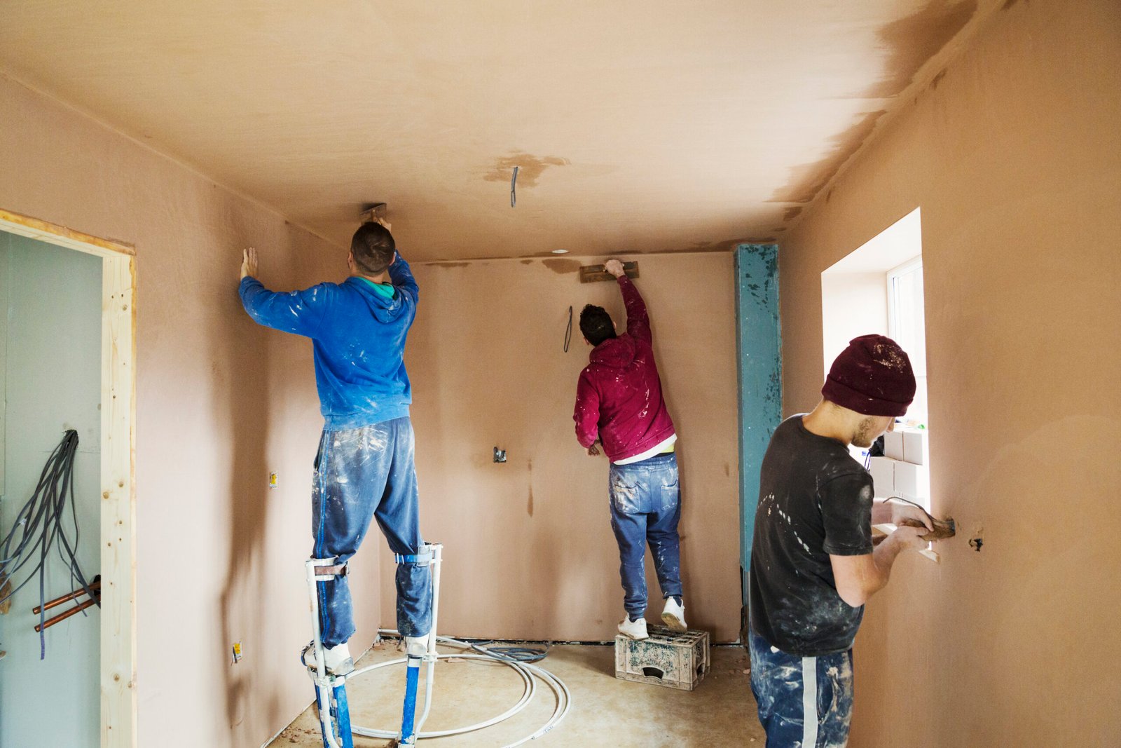 Three men, two plasterers wearing stilts smoothing fresh plaster high up on the walls of a house under construction.
